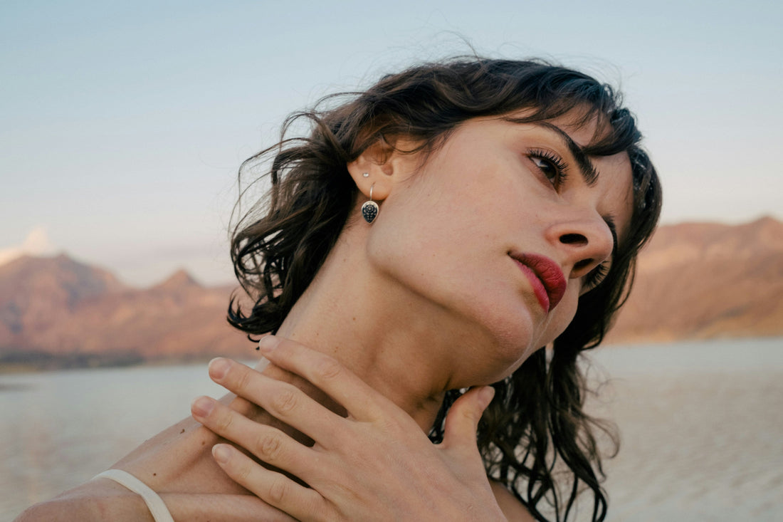 Porträt einer Frau mit geneigtem Kopf und Hand am Hals vor einer stillen Landschaft aus Wasser und Bergen.